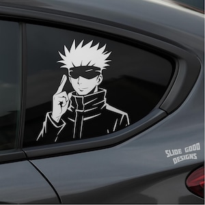 May include: White vinyl decal of an anime character with spiky hair and a blindfold, pointing upwards. The character is wearing a black jacket. The decal is on a car window. The text "SLIDE GOOD DESIGNS" is in the bottom right corner.
