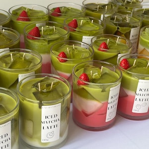 May include: A selection of candles in glass jars, designed to look like iced matcha lattes. The candles have green, white, and pink layers, with a red strawberry and clear wax cubes on top. Labels on the jars read "ICED MATCHA LATTE" and are 350g.
