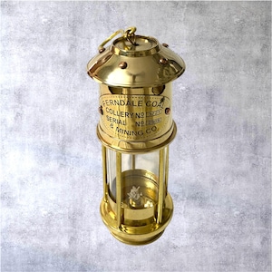 Op de afbeelding: Een messing mijnwerkerslamp met een helder glazen gedeelte. De lamp heeft de tekst "Ferndale Coal Colliery" en serienummers. Hij heeft een handvat en een voet. De lamp heeft een warme goudkleur. De lamp is ongeveer 20 cm hoog.
