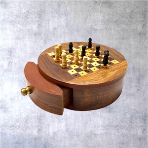 Puede incluir: Un juego de ajedrez redondo de madera con un cajón. El tablero de ajedrez está incrustado con cuadrados claros y oscuros, y las piezas de ajedrez son negras y beige. El cajón está abierto, revelando el espacio de almacenamiento. El juego tiene un pomo de latón.