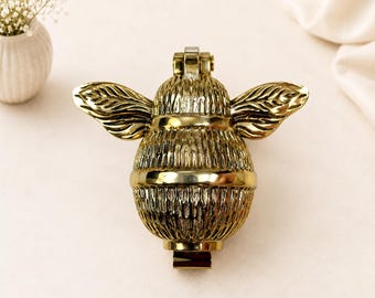 Könnte beinhalten: Ein Türklopfer aus poliertem, goldfarbenem Metall in Form einer Biene. Die Biene hat einen abgerundeten, strukturierten Körper mit horizontalen Segmenten und zwei ausgebreiteten, detaillierten Flügeln. Ein Scharniermechanismus ist oben und unten sichtbar.