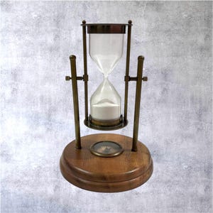 May include: An hourglass with a wooden base and brass-colored metal supports. The glass hourglass contains white sand. A compass is set into the wooden base. The hourglass is a decorative item.