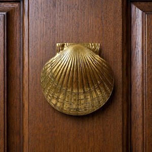 May include: A gold-toned metal door knocker shaped like a scallop shell with detailed radial ridges and a textured surface. The shell is mounted centrally on a dark brown wooden door panel featuring vertical grain and a recessed rectangular frame.