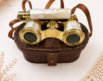 May include: Vintage-style opera glasses featuring a polished brass frame with inlaid white mother-of-pearl panels on the barrels and handle. The binoculars include a central focus knob and are resting on a dark brown leather carrying case with a strap.