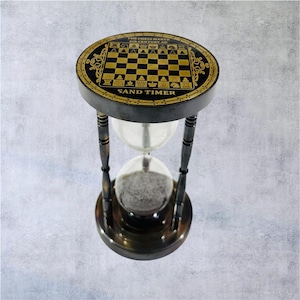 May include: A decorative sand timer featuring a chess board design. The top and base are dark brown with gold detailing. The hourglass has a clear glass centre filled with sand. Text reads "THE CHESS MAKER" and "SAND TIMER".