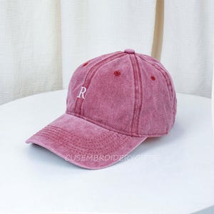 May include: A dusty rose baseball cap with a curved brim. The cap features the letter "R" embroidered in white on the front. The text "CUSEMBROIDERYGIFTS" is printed on the brim. The cap has a vintage, worn look.