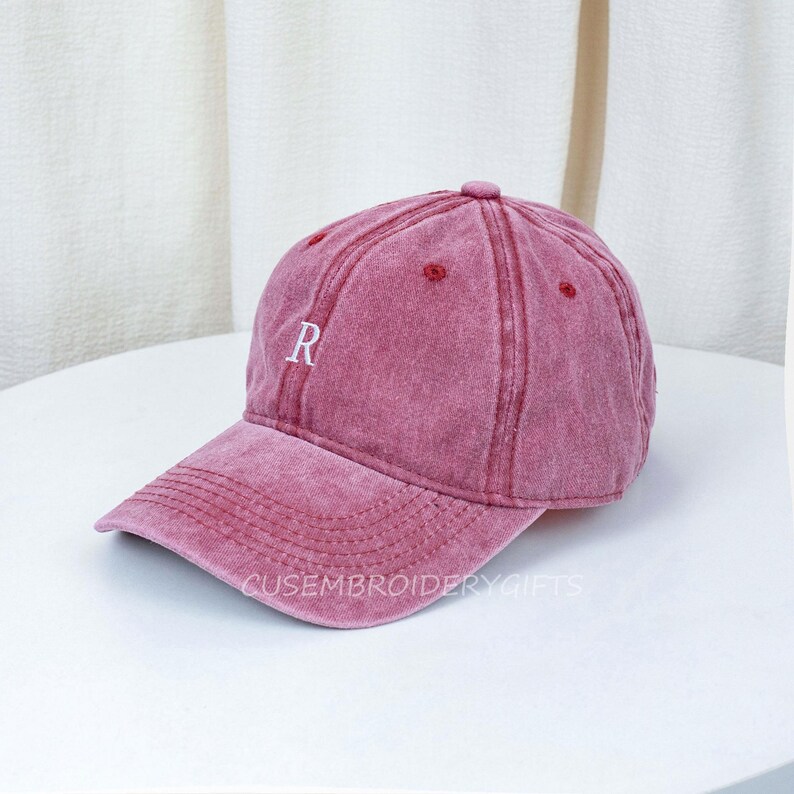 May include: A dusty rose baseball cap with a curved brim. The cap features the letter "R" embroidered in white on the front. The text "CUSEMBROIDERYGIFTS" is printed on the brim. The cap has a vintage, worn look.