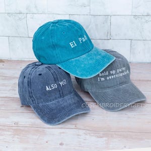 May include: Three baseball caps in shades of blue and gray are stacked on a wooden surface. The top cap is teal with "El Padre" embroidered in white. The middle cap is navy blue with "ALSO YES!" and the bottom cap is gray with "hold up partner I'm overstimulated".