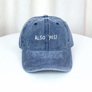 May include: A denim blue baseball cap with the embroidered text "ALSO YES!". The cap has a curved brim and a fabric strap closure. The cap is a washed denim style.