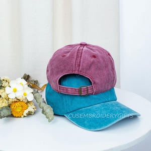 May include: Two baseball caps stacked on top of each other. The top cap is a faded maroon color, and the bottom cap is teal. The teal cap has the text "CUSEMBROIDERYGIFTS" embroidered on the brim. The caps are on a white surface.
