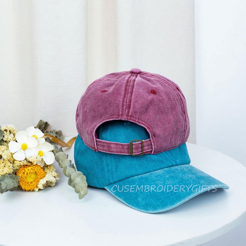 May include: Two baseball caps stacked on top of each other. The top cap is a faded maroon color, and the bottom cap is teal. The teal cap has the text "CUSEMBROIDERYGIFTS" embroidered on the brim. The caps are on a white surface.