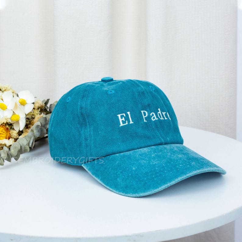 May include: A teal baseball cap with the embroidered text "El Padre" in white. The cap is made of a soft, textured fabric and features a curved brim. The cap is sitting on a white table.