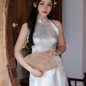 May include: A woman in a white halter dress with a floral design, holding a wooden fan. The dress features a high neckline and a flowing skirt. The website AODAIUS.COM is visible in the image.