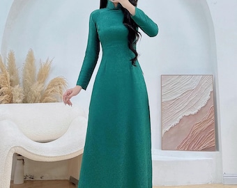 Op de afbeelding: Een lange, smaragdgroene jurk met lange mouwen en een hoge halslijn. De jurk heeft een subtiel textuurpatroon en wordt gecombineerd met een witte broek. De afbeelding bevat de tekst "AoDaiUS Brings VN Cultures to US".