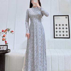 May include: A long, light grey dress with a floral pattern. The dress has long sleeves and a high neckline. The text "AODAUS.COM Brings VN Cultures to US" is visible in the upper left corner. The dress is worn with white shoes.