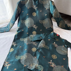 May include: Two matching teal-colored silk dresses with long sleeves and mandarin collars. The dresses feature a pattern of gold dragons, clouds, and circular designs. The dresses are displayed on a wooden hanger.