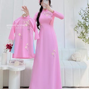 May include: Matching pink dresses with floral embroidery. One is a long, flowing gown, and the other is a shorter dress. The dresses are worn with light blue pants. The website address "AODAIUS.COM" is visible in the image.