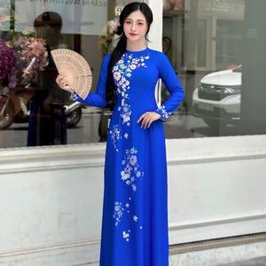 May include: A vibrant royal blue dress with a floral design, long sleeves, and a high neckline. The dress is worn by a person holding a fan. The text "AODAIUS.COM" is visible in the background. The shop's opening hours are also displayed.