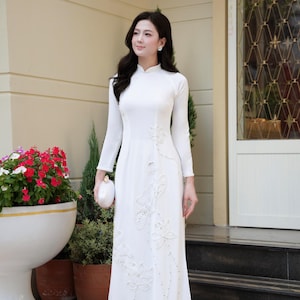 May include: A floor-length white Ao Dai featuring long sleeves, a high collar, and intricate white floral embroidery with subtle bead embellishments. The garment is paired with matching white trousers and a small white clutch purse.