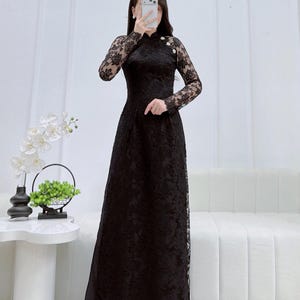 May include: A floor-length black lace ao dai featuring long sleeves, a high mandarin collar, and decorative buttons along the shoulder. The garment is crafted from intricate floral lace fabric and is paired with white pointed-toe shoes.