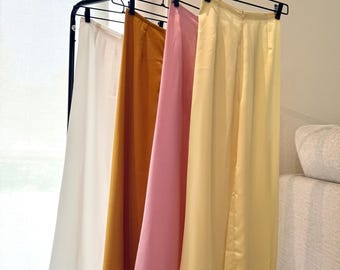 Pode incluir: Quatro pares de calças de perna larga em creme, mostarda, rosa e amarelo pálido pendurados num cabideiro preto. As calças são feitas de um tecido liso e fluido, expostas em frente a uma cortina branca.