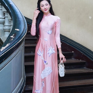 May include: A pink, long-sleeved dress with floral embroidery and a mandarin collar. The dress features white and purple floral designs and is worn with white shoes and a small white purse. The text "AoDaiUS Brings VN Cultures to US" is visible.