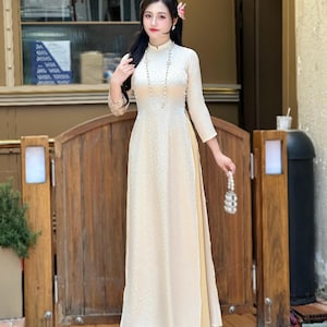 May include: A cream-colored, long-sleeved dress with a high neckline and floral embroidery. The dress is paired with wide-leg pants in a lighter shade of yellow. A pearl necklace and a small beaded purse complete the look. The outfit is suitable for formal occasions.