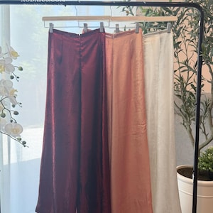 Puede incluir: Tres pantalones de pierna ancha colgados en un perchero negro. Los pantalones son de color burdeos intenso, melocotón suave y blanco roto, cada uno con una textura sutilmente brillante y un corte recto con cremallera en la parte trasera.
