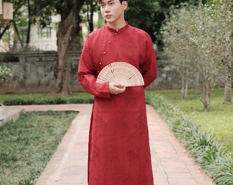 Op de afbeelding: Een persoon draagt een rode traditionele Vietnamese Áo dài, met een beige waaier. De tuniek met lange mouwen heeft een mandarijnkraag en decoratieve knopen. De outfit wordt gecombineerd met een zwarte broek. De website aodaius.com is zichtbaar.