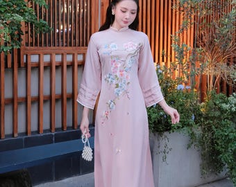 May include: A light pink ao dai dress with floral embroidery. The dress features a mandarin collar, long sleeves with pleated cuffs, and a flowing skirt. The text "AoDaiUS Brings VN Cultures to US" is visible.