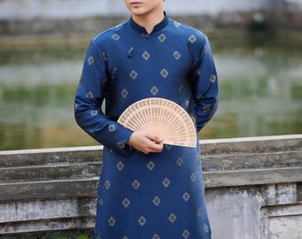 Op de afbeelding: Een donkerblauwe tuniek met lange mouwen, een mandarijnkraag en een herhalend goudkleurig geometrisch patroon, gecombineerd met een zwarte broek. Het kledingstuk heeft een knoopsluiting aan de zijkant en wordt vastgehouden met een open houten waaier.