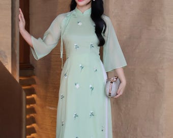 May include: A light green Vietnamese Áo dài dress with white floral embroidery. The dress has a mandarin collar and flared sleeves. The text "AODAIUS.COM Brings VN Cultures to US" is visible in the upper left corner. The dress is paired with a silver clutch.
