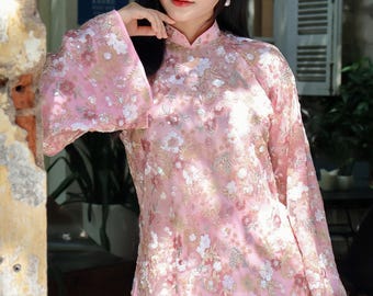 May include: A pink, long-sleeved dress with floral embroidery and bell sleeves. The dress has a mandarin collar and is adorned with white and gold embellishments. The text "AoDaiUS Brings VN Cultures to" is visible in the background.