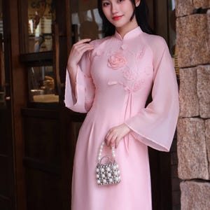 Puede incluir: Un vestido de color rosa claro con mangas largas y cuello mao, que presenta una rosa de tela tridimensional y aplicaciones de hojas en el pecho. El vestido se combina con un pequeño bolso hecho de perlas blancas y cuentas plateadas.