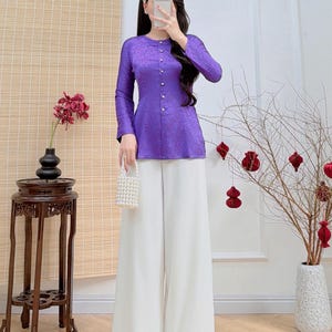 May include: A woman's outfit comprising a long-sleeved, purple top with button detailing and white wide-leg trousers. The top has a textured pattern. The outfit is paired with a small, white beaded purse. The website address AODAIUS.COM is visible.