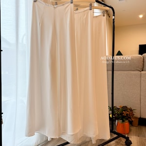May include: White, flowing pants hanging on a black clothing rack. The pants are made of a lightweight fabric and have a high waist. The text "AODAIUS.COM Brings VN Cultures to US" is visible in the image.