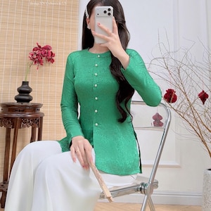 Puede incluir: Una blusa verde de manga larga con estampado floral y botones de perlas, combinada con pantalones blancos de pernera ancha. La blusa tiene cuello mandarín y abertura lateral. La dirección del sitio web AODAIUS.COM es visible.