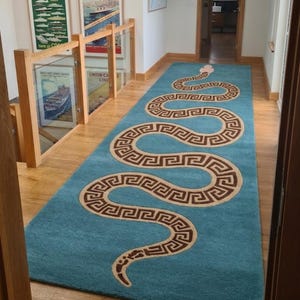 May include: A long rectangular runner rug in a vibrant turquoise blue colour. The rug features a large, winding snake design that spans the entire length of the runner. The snake has a light-coloured head and a body patterned with brown Greek key motifs.