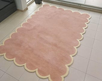 May include: A large, rectangular rug with a scalloped edge. The rug has a soft, pink center and a cream-colored border. The rug is placed on a tiled floor, and a plant in a black pot is visible in the background.