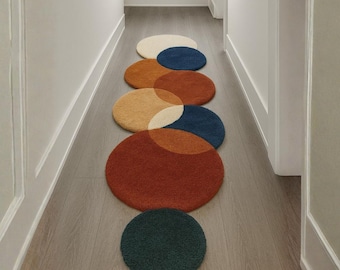 Puede incluir: Una alfombra de pasillo compuesta por alfombras circulares superpuestas de varios colores. Las alfombras son en tonos crema, azul, naranja y óxido, con una alfombra verde oscuro al final. El pasillo tiene suelo de madera clara y molduras blancas.