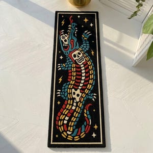 Könnte beinhalten: Ein rechteckiger schwarzer Teppich mit einer farbenfrohen Illustration eines Skelett-Alligators. Der Alligator hat einen blauen Kopf, eine rot-gelbe Wirbelsäule und einen weißen Schädel. Der Teppich ist mit einem goldenen Rand eingefasst und hat Sterne und Blitze.