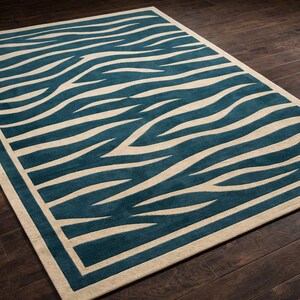 Puede incluir: Una alfombra rectangular con un diseño de rayas de cebra en color turquesa y crema. La alfombra tiene un borde crema y está colocada sobre un suelo de madera oscura. El diseño presenta rayas fluidas en forma de onda.