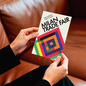 Könnte beinhalten: Eine rechteckige Werbekarte für die Mailänder Messe, die von einer Person gehalten wird. Die Karte zeigt den Text "MILAN TRADE FAIR" und ein farbenfrohes geometrisches Design. Die Karte enthält auch den Text "International Trade Centre".