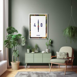 May include: A framed print with a white border featuring a stylized illustration of a knife with the numbers "4 0" and the word "COMMANDO". The background of the print has a camouflage pattern. The print is hanging on a green wall.