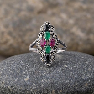 Peut inclure: Une bague en argent au design orné, ornée de pierres précieuses ovales. La bague présente un groupe central de pierres vertes, rouges et noires, entouré de petits accents étincelants. Posée sur une surface en pierre grise.