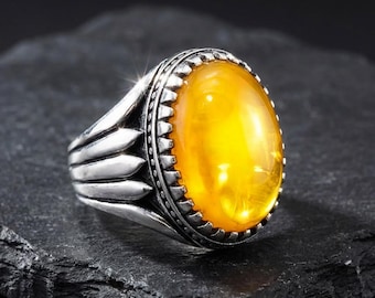 Könnte beinhalten: Silberring mit einem großen, ovalen, bernsteinfarbenen Edelstein. Der Ring hat ein detailliertes, strukturiertes Band mit einem dekorativen Rand um den Stein. Der Edelstein ist poliert und reflektiert Licht, vor dunklem Hintergrund.