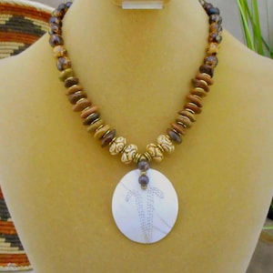 May include: A handcrafted necklace with a large, round, white pendant featuring a dotted design. The necklace has a beaded strand with brown, tan, and gold-coloured beads. The necklace is displayed on a mannequin.