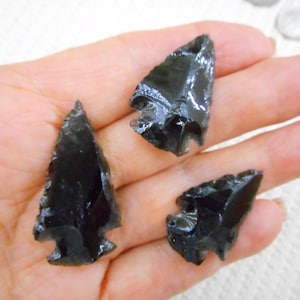 May include: Three black obsidian arrowheads with sharp edges are displayed in a hand. The arrowheads have a glossy, reflective surface and are triangular in shape. They are likely handmade and are a dark, opaque color.