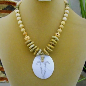 May include: A beige beaded necklace with a large white pendant. The necklace features round and disc-shaped beads in shades of beige and brown, with gold-coloured accents. The pendant has a dotted design.