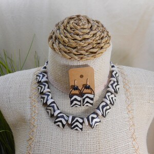 May include: A necklace and earring set displayed on a mannequin. The necklace and earrings feature black and white geometric patterns. The earrings are rectangular with black hoops. The necklace is made of square beads.